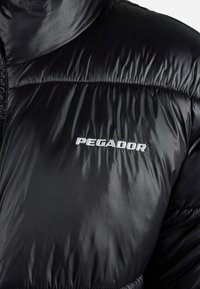 Svart, skinnende dunjakke med hvit brodert "PEGADOR"-logo på brystet, som viser detaljert stofftekstur og søm.