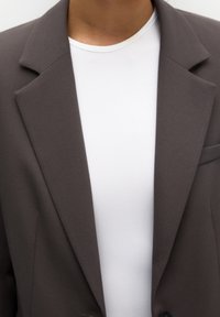 Blazer marron sur mesure avec une texture lisse, présentant un revers cranté et une fermeture à un bouton, porté sur un t-shirt blanc.