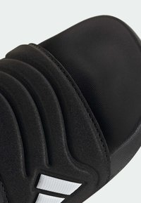 Černá pantofle na klínu s texturovaným páskem se zvýrazněnými liniemi a bílými proužky jako akcentem. Materiál vypadá jako syntetický s hladkým povrchem.