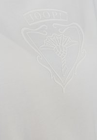 Hvid bomulds T-shirt med et hævet, præget logodesign med et dekorativt skjold og blomstermønster i midten.