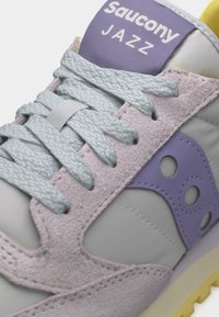 Gros plan sur une chaussure Saucony Jazz avec des lacets bleu clair, des détails en daim lavande et un logo violet sur un tissu gris clair.