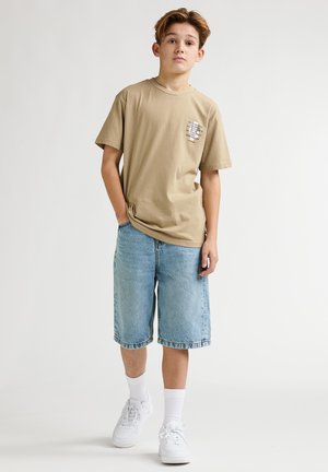 Camiseta de algodón beige con un pequeño diseño impreso en el pecho, combinada con pantalones cortos de mezclilla azul claro que llegan hasta las rodillas y zapatillas blancas.