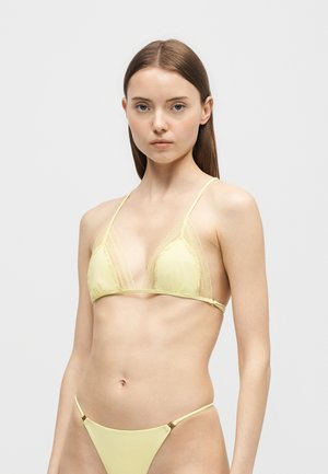 Jeune femme aux cheveux bruns et raides portant un bikini jaune clair avec une bordure en dentelle sur un fond clair uni.