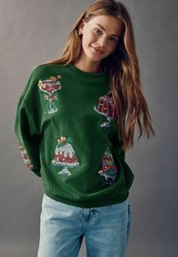 Hanorac verde cu desene colorate din paiete care reprezintă deserturi și băuturi festive. Croială lejeră, gât rotund și material moale.