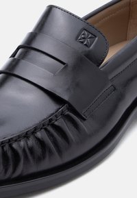 Mocassin en cuir noir avec bride à empiècement, surpiqûres détaillées et logo embossé sur la partie supérieure, semelle lisse visible sur fond blanc.