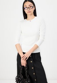 Vit ribbade långärmade topp med vågiga kanter, kombinerad med en svart kjol med knappar fram. Håller en svart blommig väska med samlade handtag.