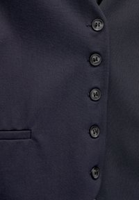 Granatowa kamizelka wykonana z gładkiego materiału, z sześcioma czarnymi guzikami ułożonymi w pionową linię oraz boczną kieszenią po lewej stronie. Widoczna faktura powierzchni.