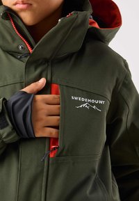 Grön vattentät jacka med röda detaljer, utrustad med hög krage, dragkedjeficka och "SWEDEMOUNT"-logotyp. Mjuka muddarna har bra grepp och tyget är slitstarkt.