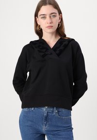 Melnā kapucē ar V veida izgriezumu, kurai ir teksturēts melns raksts uz kapuces. Saskaņots ar ziliem džinsiem. Gluda auduma, brīvi pieguļošs. 
