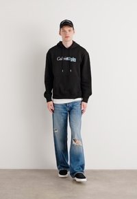 Black hoodie with blue "Calvin Klein" logo, light blue ripped jeans, black shoes, and a black cap. Plain background, standing model.