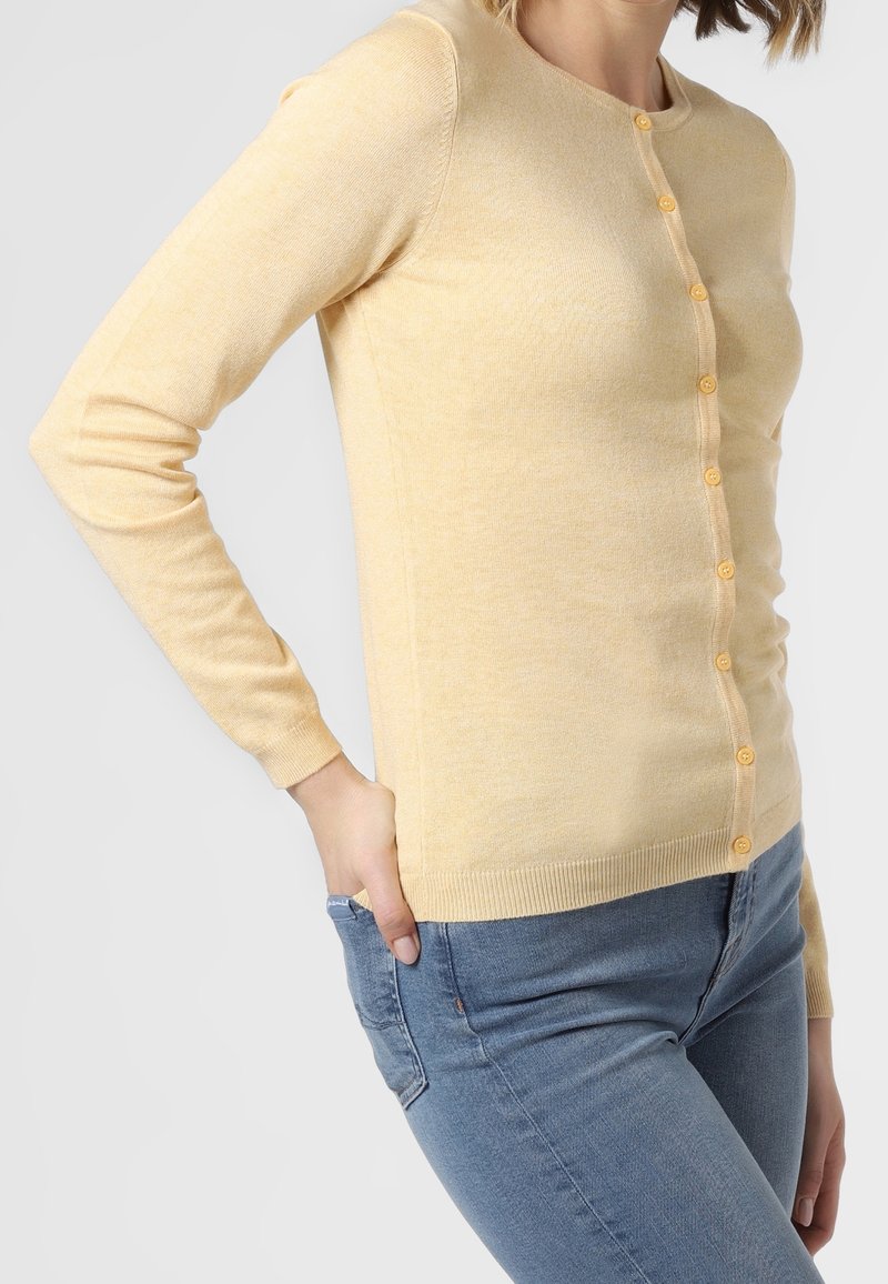 Femme portant un cardigan ajusté jaune pâle boutonné et un jean bleu, une main dans la poche, sur un fond clair uni.
