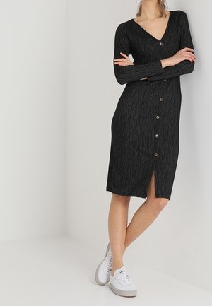 Kvinde iført en sort, langærmet kjole med knapper og frontspalte, stående med armene krydsede og hvide sneakers på en ensfarvet baggrund.