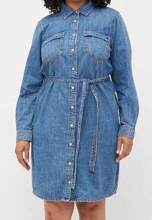 Kvinde iført en blå denimskjortekjole med lange ærmer, foran knapper, to brystlommer og et bindebælte i taljen.