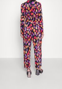 Kobieta ubrana w kombinezon z długim rękawem w intensywny kwiatowy wzór w kolorach jasno pomarańczowym, czerwonym, kremowym i fioletowym na czarnym tle, do tego sandały na słupku.