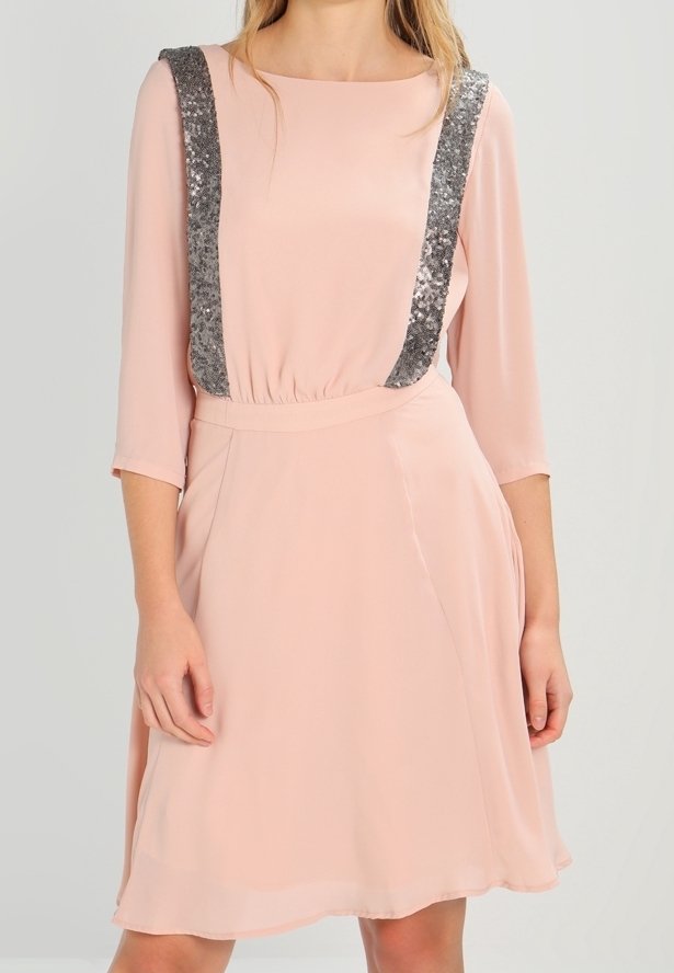 Femme portant une robe rose pâle arrivant au genou, avec des manches trois-quarts et des bretelles argentées à paillettes attachées à la taille.