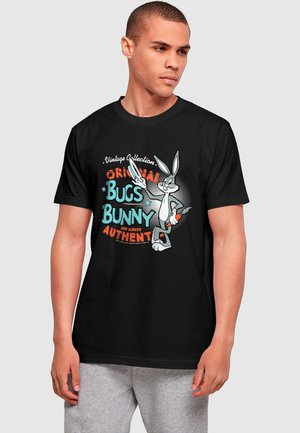 Mladý muž nosí čierne tričko s vintage grafikou Bugs Bunnyho a textom "Original Bugs Bunny and Always Authentic" v modrej a oranžovej farbe.