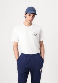 Baltā kokvilnas T-kreklā ir tumši zilā krāsā izdrukāts "Tommy Hilfiger 1985", savienots ar tumši zilām elastīgām svīterbiksēm, kam priekšpusē ir baltā krāsā izdrukāts "NY". Zils cepures.