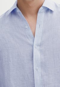 Lichtblauwe, geknoopte shirt met een gestreepte textuur en knopen aan de voorkant. De stof lijkt lichtgewicht en ademend.