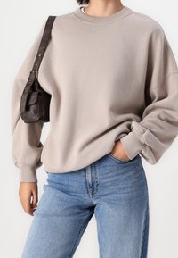 Osoba v béžovém oversized svetru, modrých džínách, s malou tmavě hnědou koženou kabelkou na rameni a jednou rukou v kapse.