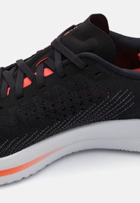 Zapatilla deportiva negra y roja con parte superior de punto, cordones negros y suela blanca con un detalle de raya roja.