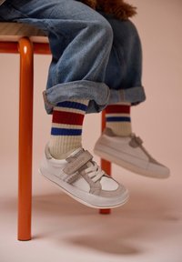 Baskets blanches avec des accents gris et une sangle Velcro, portées avec un jean en denim retroussé et des chaussettes en maille multicolores présentant des rayures rouges et bleues.
