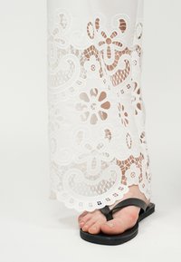 Πόδι που φοράει μαύρο σανδάλι με λουράκι στα δάχτυλα κάτω από λευκό ένδυμα δαντέλας με κεντήματα ανθολογίας και στροβιλισμού.