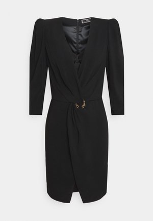 Robe noire longueur genoux avec manches longues, décolleté en V profond, détail froncé à l'avant et ornement en chaîne dorée à la taille.