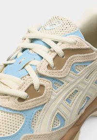 Zapatilla deportiva con paneles de malla y ante en beige, azul claro y marrón, con cordones blancos y suela texturizada.