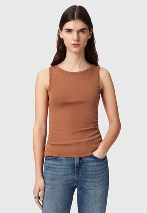 Kobieta z długimi ciemnymi włosami, ubrana w bezrękawnik w kolorze brązowym i niebieskie dżinsy, stoi z jedną ręką w kieszeni na jednolitym tle.