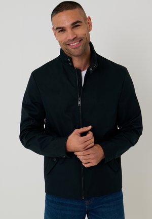 Uśmiechnięty mężczyzna z krótkimi włosami, ubrany w czarną kurtkę na zamek i niebieskie dżinsy, stojący na jasnym, jednolitym tle.