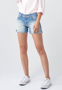 Femme portant un short en denim bleu clair, des baskets blanches et une chemise rose clair à manches retroussées, debout devant un fond uni.