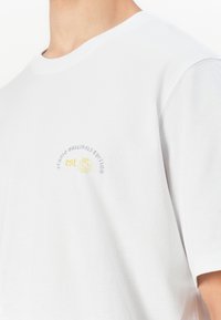 Hvit t-skjorte med lite grått og gult logo som leser "Studio Originals Edition est. 99" på brystet, båret av en person som er klippet ved halsen.