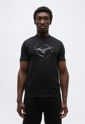 Άνδρας που φοράει μαύρο κοντομάνικο μπλουζάκι με κυκλικό λευκό αφηρημένο λογότυπο, στέκεται μπροστά σε απλό ανοιχτό γκρι φόντο.