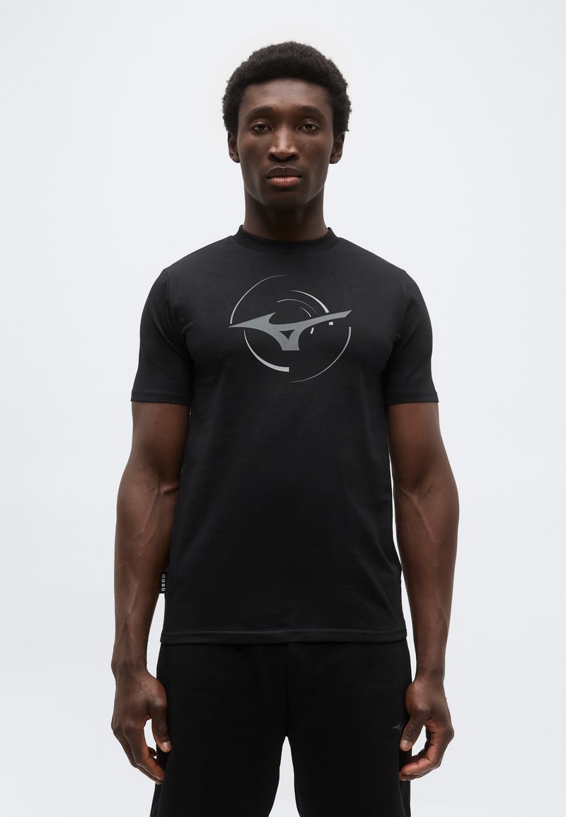 Άνδρας που φοράει μαύρο κοντομάνικο μπλουζάκι με κυκλικό λευκό αφηρημένο λογότυπο, στέκεται μπροστά σε απλό ανοιχτό γκρι φόντο.