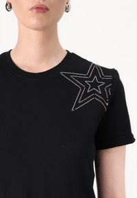 Black cotton t-shirt featuring rolled sleeves and a rhinestone star design on the left shoulder. Smooth texture and fitted shape.