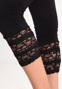 Svarta leggings med rynkdesign vid låret. Slutet pryds av utsökt svart spets med ett blommönster, vilket skapar en texturerad kontrast.