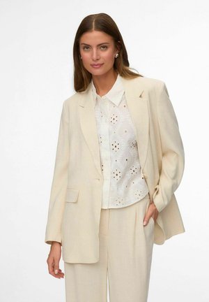 Kobieta o długich brązowych włosach, ubrana w kremową marynarkę, białą bluzkę z ażurowym wzorem i dopasowane kremowe spodnie, pozująca z jedną ręką w kieszeni.