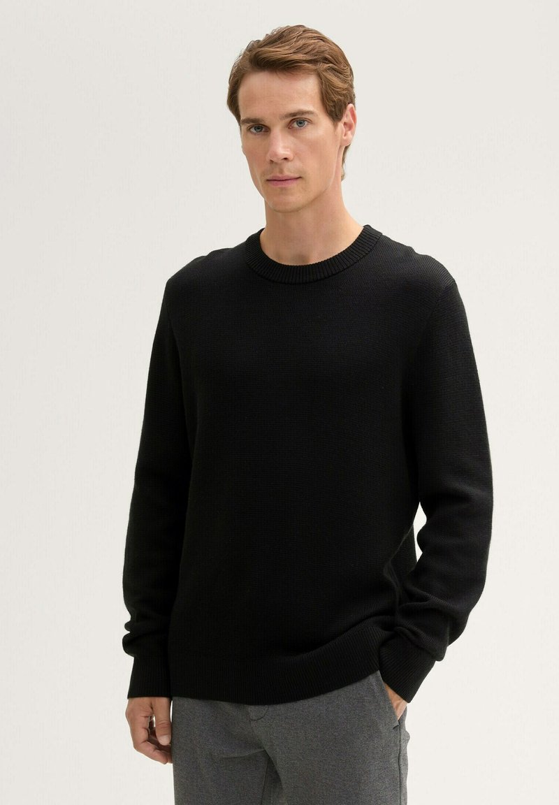 Schwarzer gestrickter Pullover mit rundem Ausschnitt und gerippten Bündchen. Der Stoff wirkt weich und strukturiert und wird in einem einfachen, taillierten Design gezeigt.