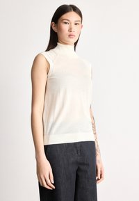 Ujjatlan kötött felső világos bézs színben, magas nyakkal, sima textúrával és kontrasztos bordázott szegéllyel. Sötét farmernadrággal viselve.