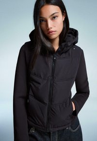Zwarte puffer vest met capuchon, ritssluiting aan de voorkant en opgestikte zakken. Zachte stof met een doorgestikt ontwerp en gladde mouwen, gecombineerd met donkere jeans.