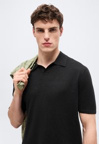 Jeune homme aux cheveux bruns courts et bouclés portant un polo noir à manches courtes, tenant une veste à carreaux verts sur l'épaule, sur un fond clair uni.