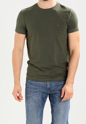 Mężczyzna ubrany w oliwkowozielony t-shirt z krótkim rękawem i okrągłym dekoltem oraz niebieskie dżinsy, stojący na jednolitym jasnym tle.