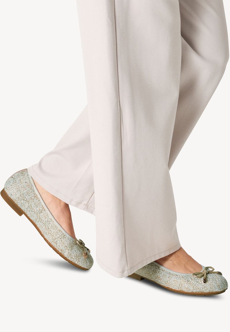 Scarpe basse con una tomaia blu chiaro texture, una piccola accentatura a fiocco e una suola marrone, abbinate a pantaloni a gamba larga di colore chiaro.