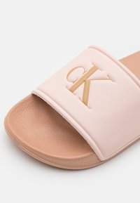 Ružová šľapka s hladkým matným povrchom a vyvýšeným zlatým logom "CK". Textúrovaná béžová stielka pre lepšiu priľnavosť a pohodlie.