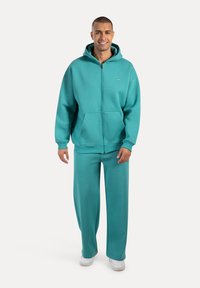 Turkusowa bluza z zamkiem z przodu i kieszenią oraz dopasowane spodnie, wykonane z miękkiego materiału. Model nosi białe sneakersy.