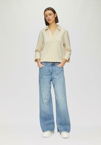 Kremowa bluzka z długim rękawem w kształcie litery V i szerokimi rękawami, zestawiona z jasnoniebieskimi szerokimi spodniami jeansowymi i białymi trampkami. Prosty, casualowy design.