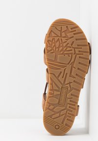 Sandale marron avec une semelle en caoutchouc texturée présentant un motif de feuilles et un logo de marque. Les lanières offrent un maintien sécurisé du pied.
