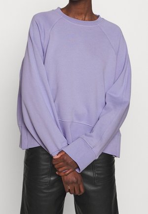 Persoon draagt een pastelpaars sweatshirt met lange mouwen en geribbelde manchetten, en zwarte leren broek, handen voor zich gevouwen.