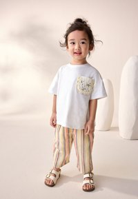 Fehér rövid ujjú póló sárga hullámos szegélyű, virágmintás hímzett zsebbel, színes csíkos, felhajtott nadrággal és szandállal párosítva.