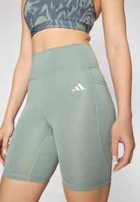 Lys grønne treningsshorts laget av stretchy stoff, med høy midje og sidelommer, og en hvit logo på låret.